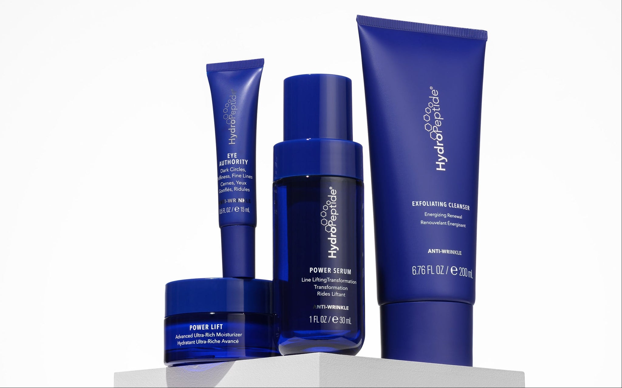 A set of blue peptide based products from the brand Hydropeptide, including a tall squeeze tube of exfoliating cleanser, a cylindrical bottle of peptide line lifting serum, a small jar of ultra rich moisturizer, and a tall rectangular tube of eye cream fo dark circles, fine lines, and puffiness, arranged on a white pedestal against a clean white background.